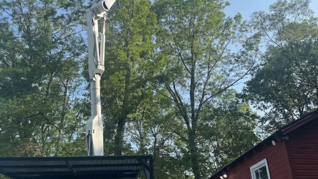 White bucket truck with elevated boom working near tall trees beside a small red building