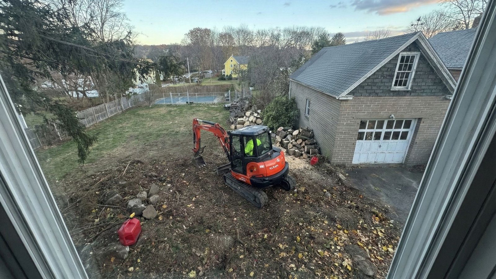 Window view of a compact excavator clearing tree debris at a residential property in Seymour Connecticut